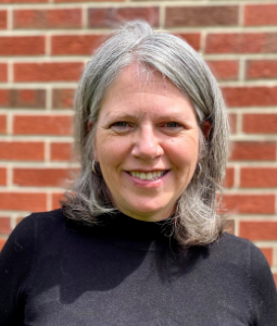 A woman with grey hair and black shirt smiles at the camera in front of a brick wall