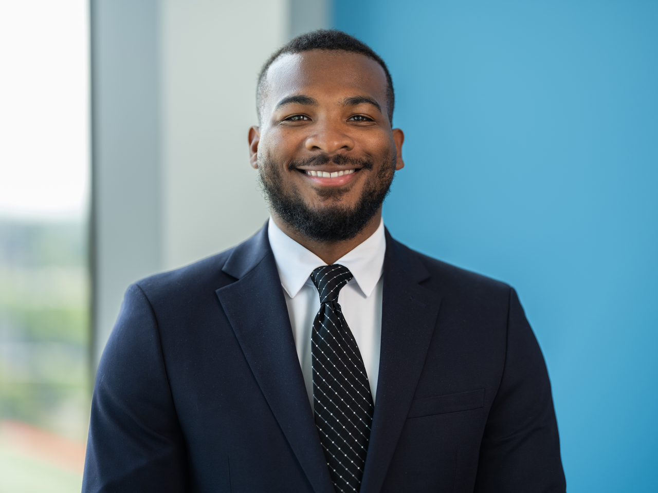 A black man wearing a navy suit, dark tie, and white shirt smiles