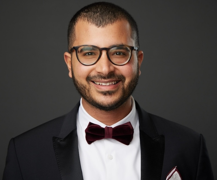 A man with a medium brown complexion wears glasses, a tuxedo, and smiles into the camera.