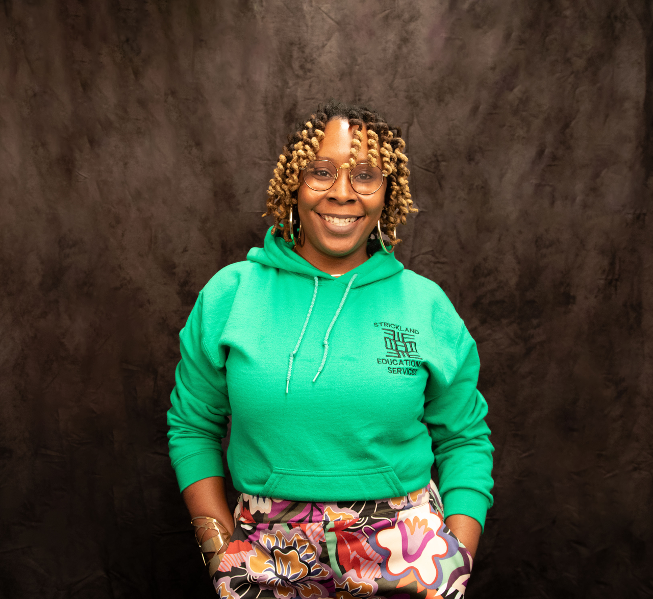 A black woman with curly hair wears a green sweatshirt smiling at the camera in front of a dark backdrop