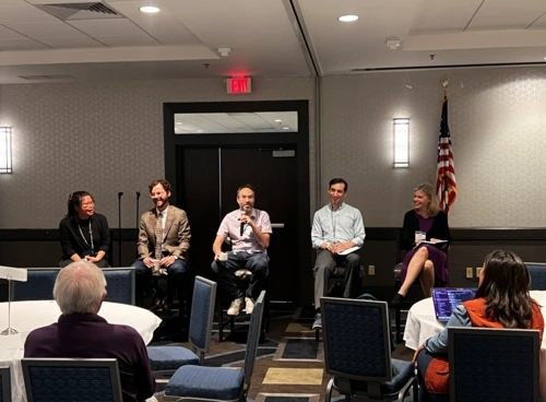 Ryan Baker sits on a stool, speaking into a microphone, among other panelists in front of an audience at ICQE.
