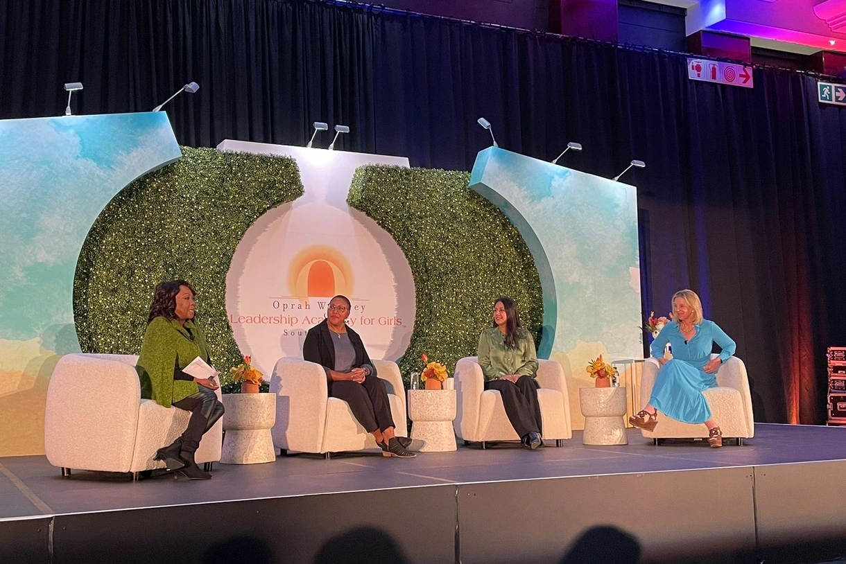 Four women sit in upholstered chairs on a black stage, a backdrop set piece behind them of walls painted blue with white clouds and greenery in the shape of an “O” around the Oprah Winfrey Leadership Academy for Girls logo.