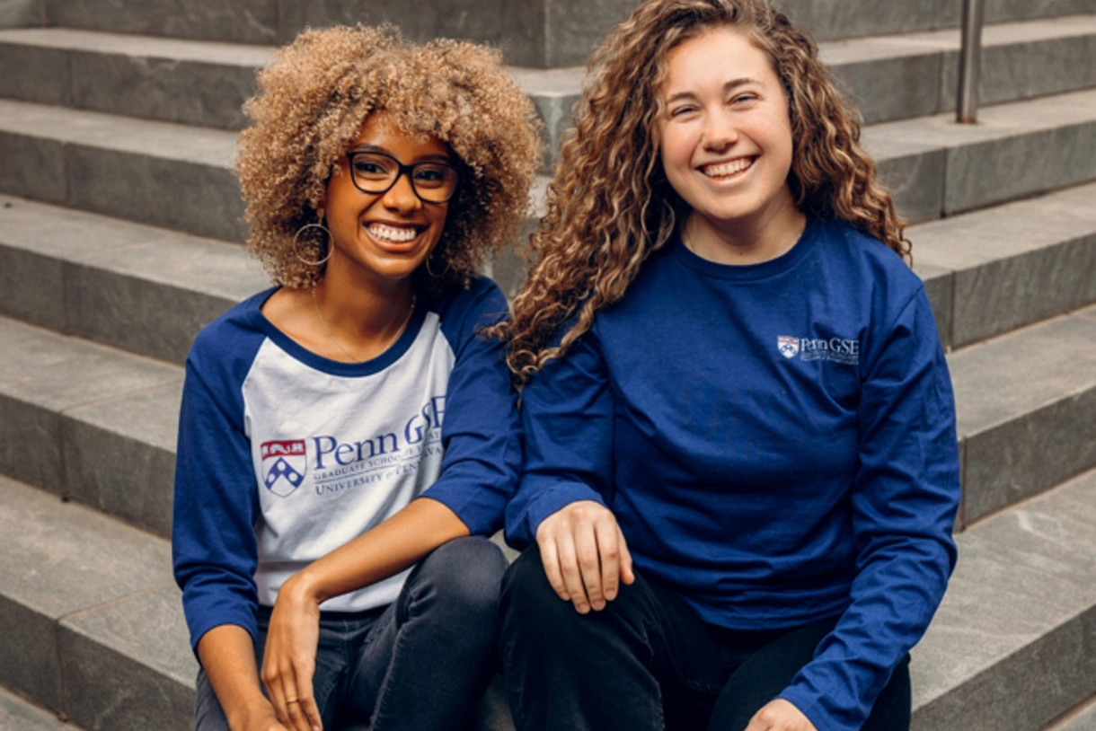 Two graduate students sit on the stairs outside Penn GSE in blue GSE long-sleeved t-shirts.