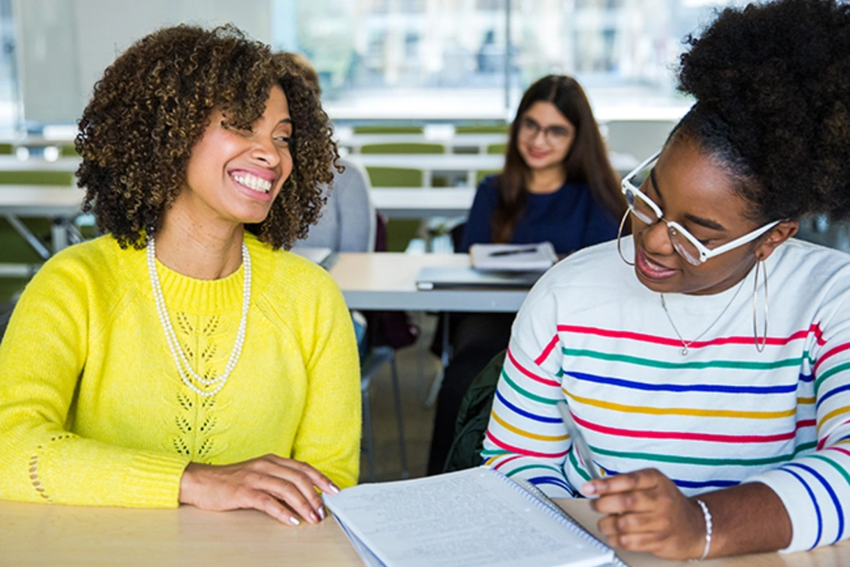 A professor and student wearing bright colors sit at a desk in a classroom with smiles on their faces, discussing academic material.