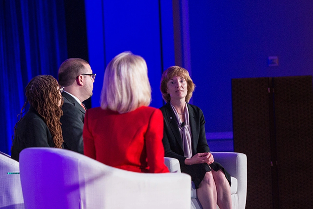 Four individuals in professional attire are seated in front of a blue curtain on a stage and engaged in conversation.