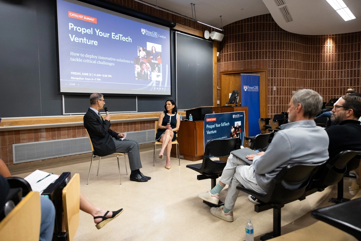 Penn GSE Dean Katharine Strunk, seated in a chair in front of an audience in a lecture hall, speaks into a microphone as Vice Dean of Innovative Programs and Partnerships Michael Golden listens, seated in a chair next to her. A screen behind them reads "Catalyst Summit: Propel Your EdTech Venture | How to deploy innovative solutions to tackle critical challenges."