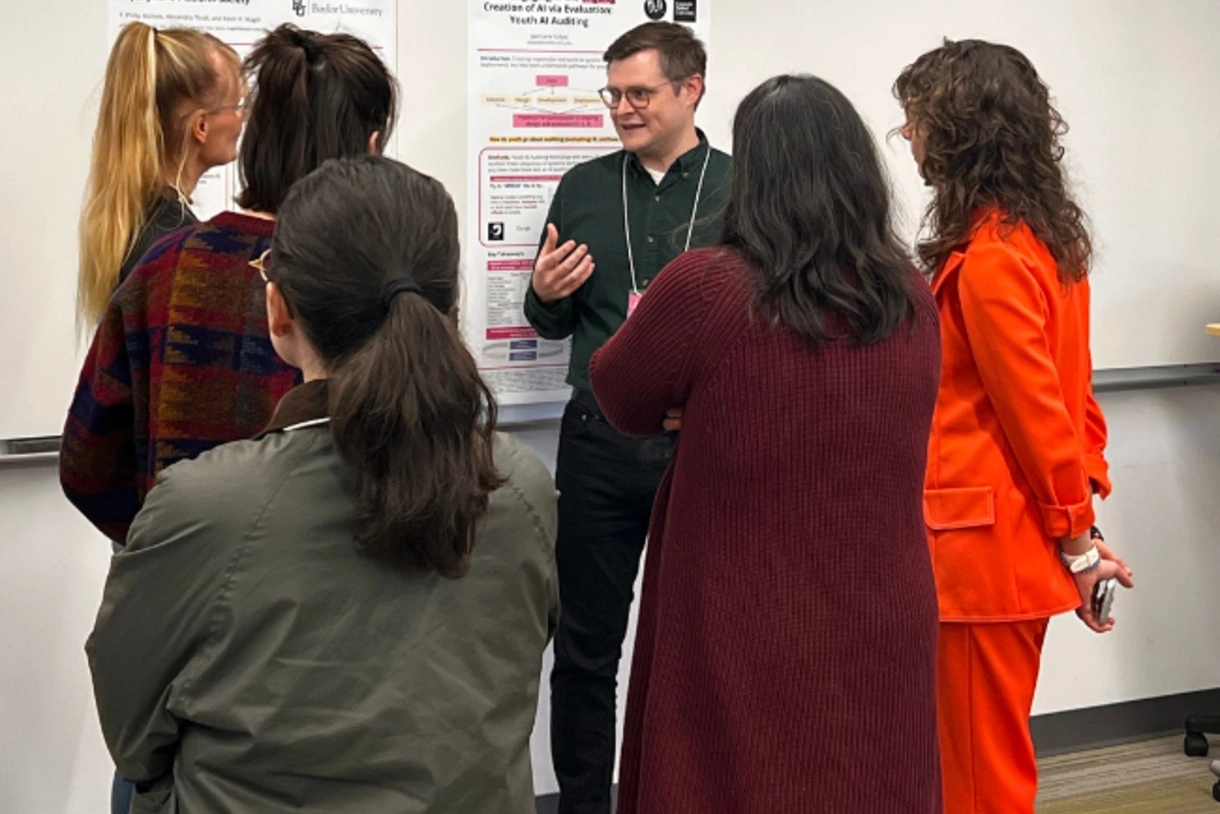 A scholar presents on a research poster hung on the wall behind them to several workshop attendees.