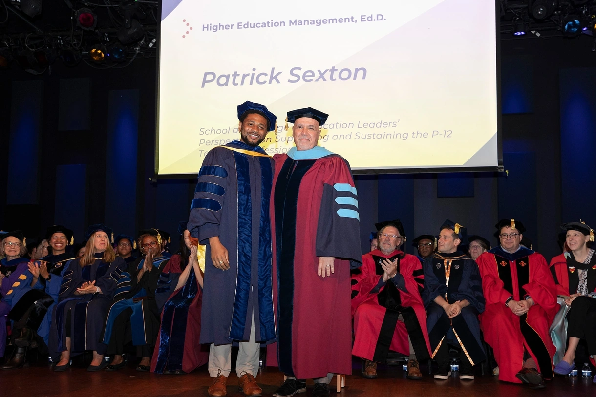Patrick Sexton poses with Assistant Professor Damani White-Lewis in their Commencement robes on stage with Penn GSE faculty in robes seated behind them and Patrick's name and degree on a screen behind them