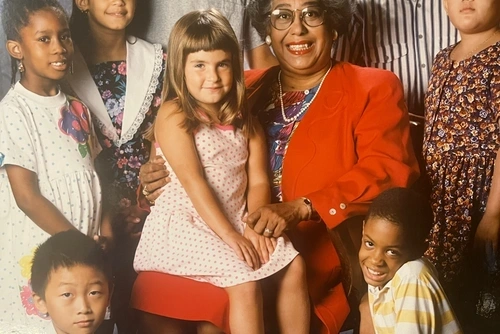 Dr. Constance E. Clayton sits in a chair wearing a red skirt and blazer over a floral shirt, surrounded by a multiracial group of children from the Philadelphia School District, during her term as Philadelphia's superintendent in the 1980s and early 90s, a young girl seated on her lap, all smiling at the camera.