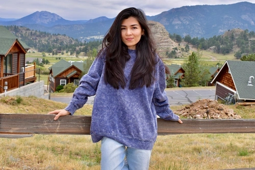Yasmeen wears a purple sweatshirt and jeans as she leans on a fence with the RockyMountains behind her.