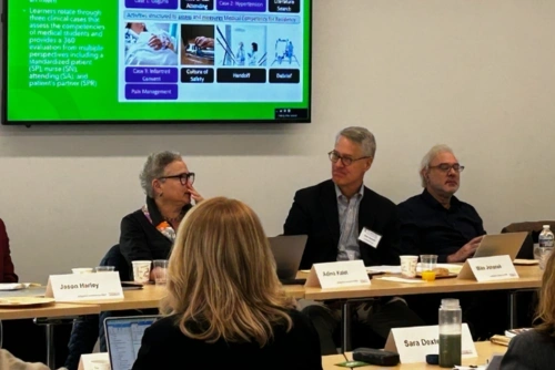 Image of 3 members of a panel in front of a green powerpoint slide with 2 members of the audience visible.