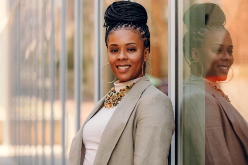 Ericka S. Weathers, with a warm smile, stands confidently, her braided hair elegantly styled into an updo, complemented by large hoop earrings, a vibrant beaded necklace, and a chic beige blazer, reflected against a glass surface.
