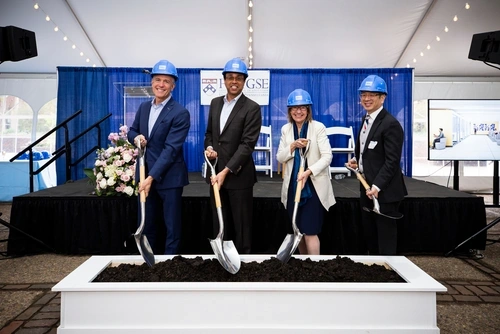 Four individuals standing next to each other and facing frontwards are wearing blue-colored hard hats and putting shovels into a pile of dirt