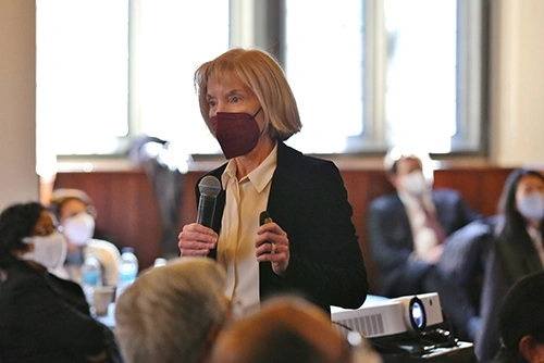A professor in a white shirt and black blazer holding a microphone and speaking to an audience.