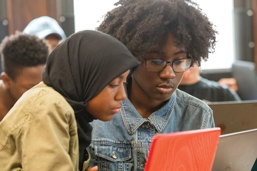 wo adolescent students in a classroom look down at a laptop screen