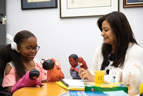 An elementary school student sitting at a table in an office looks down at two puppets held in her hands, and across from her is a smiling woman holding two other puppets. A notebook and folders are on the table.