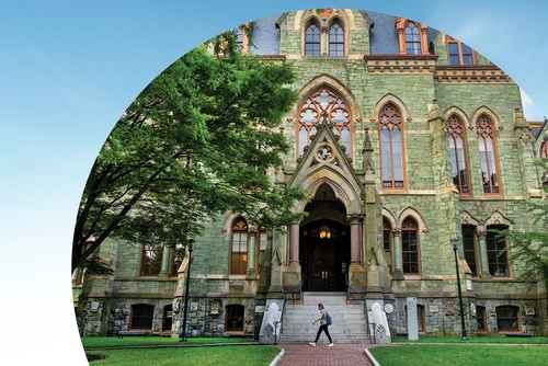 A student walks in front of College Hall building on the Penn campus., towards a leafy green tree. The photo is cropped with a semicircular edge against a blue-sky background.