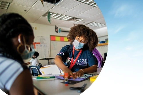 An instructor wearing a mask and a Penn GSE T-shirt is and engaged in conversation with a school student as they work on a project on a table. The photo is cropped with a semicircular edge against a blue-sky background.