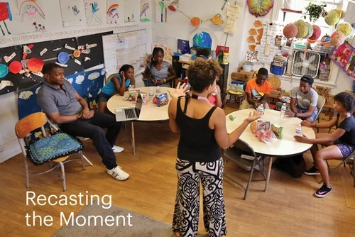 Elementary school students sit at round tables in a classroom. Behind them are colorful decorations, classroom projects, and school supplies. A woman stands at the front of the room and a man sits to the left. A headline on the image reads, “Recasting the Moment.”