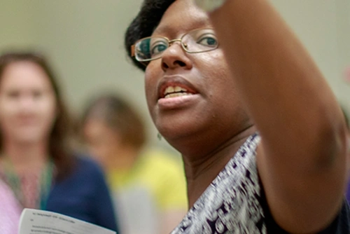 Adrienne Whaley holds notes in her one hand points towards something behind her as she teaches a room of individuals in the background.