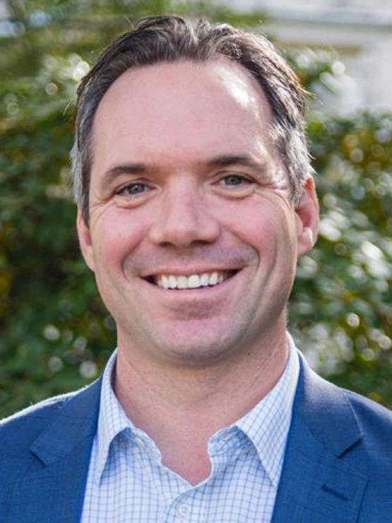 A headshot of Eric Hartman, smiling in front of a green, leafy tree