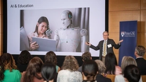 A speaker delivers a talk about AI & education in front of a screen featuring a robot teaching a student on an iPad