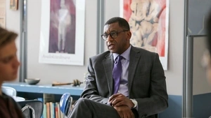 Dr. Howard Stevenson is seated in front of a high school classroom, facing students who are out of focus in the foreground. He appears relaxed and casual, resting his hands in his lap. He is wearing a grey suit with a purple tie and is listening to one of the students off-camera who is sharing an experience.