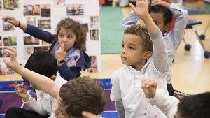 Children raise their hands while sitting on a mat in Kindergarten classroom