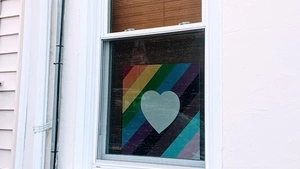 A sign with a heart over a pride flag hangs in the window of a house. 