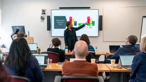 Adult Executive Doctorate in Education Management students seated at rows of tables in the foreground as an instructor stands in front of them pointing to a colorful financial aid graph on a screen titled “Need Varies Based on Cost.”