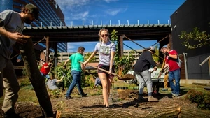 A young woman shoveling dirt at the Penn Park Orchard with other volunteers working and Patient Pavilion in the background.