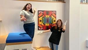 Two young women pose in front of a multicolored abstract art piece along the staircase in the Penn GSE lobby.