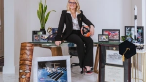 Lauren in a suit sitting on a desk surrounded by awards, framed diplomas, a model rocket, plants, space images, and sports memorabilia.