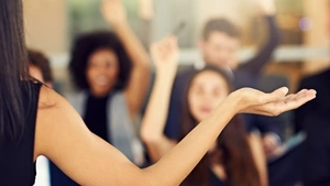 A woman instructor’s arm outstretched in the “I don’t know” gesture while people seated in front of her raise their hands with questions.
