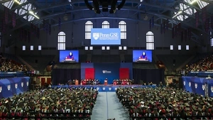 Penn GSE's graduation ceremony in the Palestra with rows of students in caps and gowns facing a stage while Angela Duckworth speaks and is visible on two large screens at the front. 