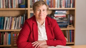 Karen Weaver smiles, wearing a red blazer and seated at a desk in front a full bookshelf