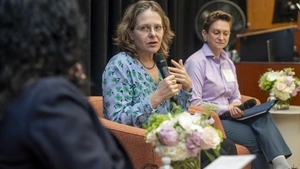 Three women sit in armchairs on a panel. The woman in the middle speaks with a microphone. 
