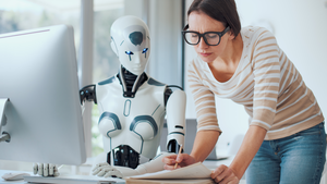 A student writing with pen an paper stands beside an AI robot typing at a computer as the two work together.